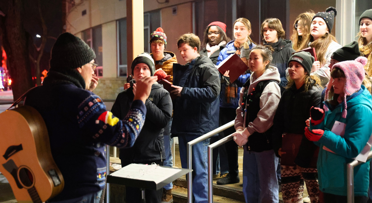 A teacher wearing a guitar on his back conducts students standing outside in a group